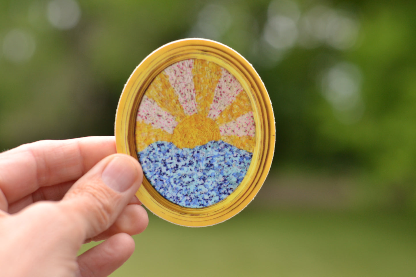 Hand holding a round, hand-painted sticker with a sun design against a blurred green background